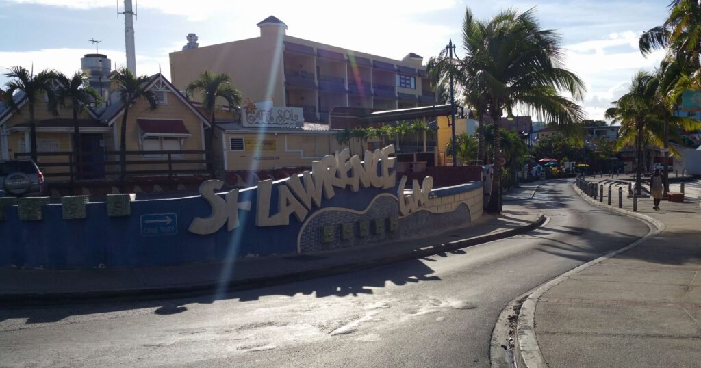 Entrance sign of St. Lawrence Gap with a street view, palm trees, and a clear blue sky in Barbados