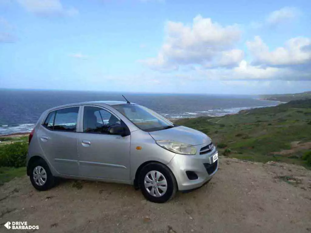 Silver economy car parked on a cliff with a panoramic view of the East Coast of Barbados in the background