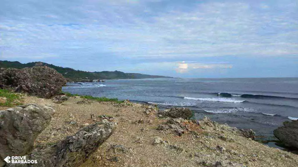 Surfers catching waves at the famous Soup Bowl surfing spot in Bathsheba, Barbados, with rugged rocks in the foreground