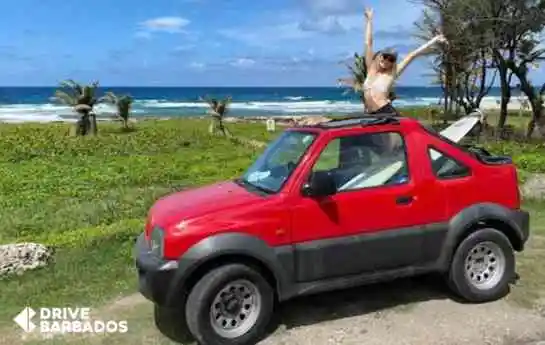 Female surfer standing up in parked red Suzuki jeep at East Coast beach, Barbados